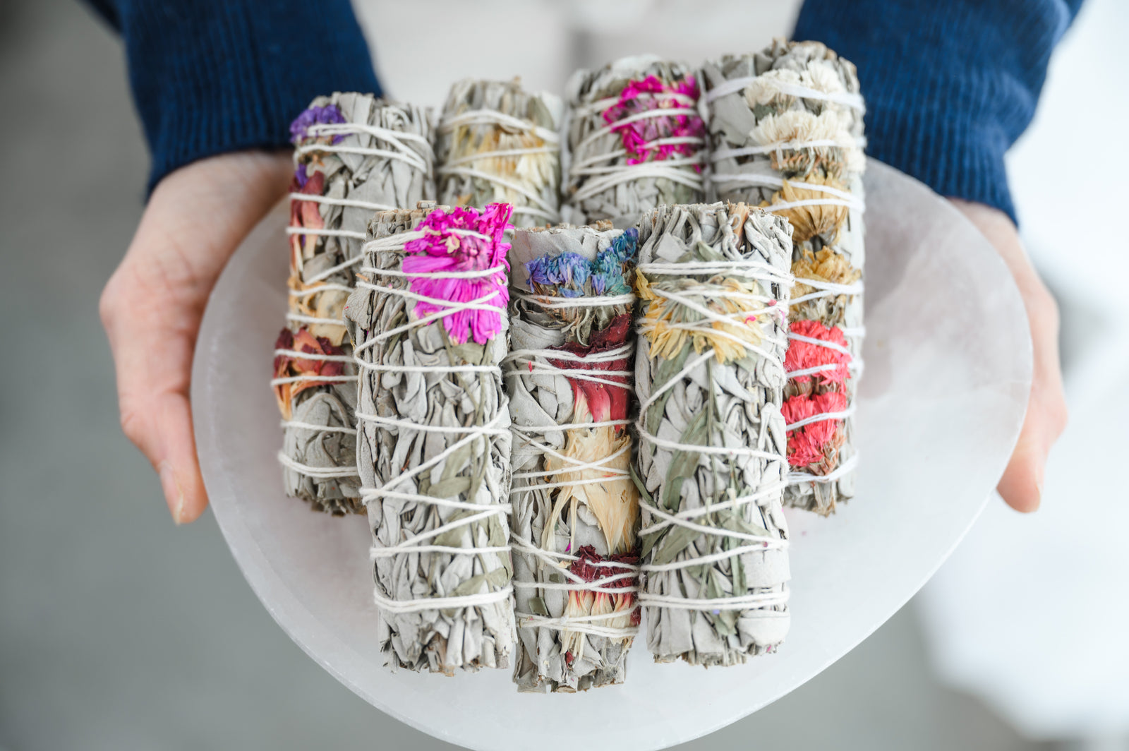 A person holding a collection of sage bundles, each bundle is wrapped with dry leaves and colorful wildflowers.